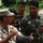 The image shows a group of military personnel engaged in what appears to be a briefing or discussion outdoors. A Western soldier wearing desert camouflage (MARPAT pattern) and a cover cap is speaking and gesturing to several soldiers wearing woodland camouflage uniforms, likely of Asian origin. The setting appears to be a garden or outdoor area with green vegetation in the background.