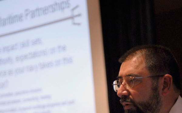 A Canadian naval officer is presenting at what appears to be an Enlisted Symposium, speaking at a microphone. Behind him is a projected presentation slide titled 'Global Maritime Partnerships' with bullet points discussing skill sets and workforce topics. He is wearing a white naval uniform with Canadian insignia and service ribbons.