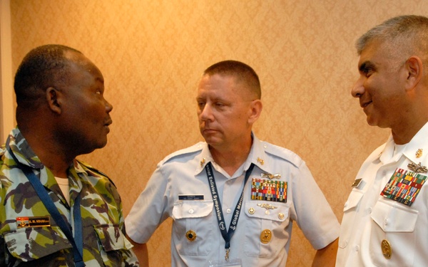 Three military personnel in different uniforms are engaged in conversation in what appears to be a formal indoor venue. The individuals represent different branches or nations, as evidenced by their distinct uniforms. One person is holding what appears to be a dark-colored item, possibly a hat or equipment.