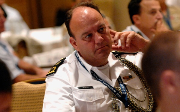 The image shows a group of military personnel seated in what appears to be a conference or symposium setting. A central figure in a white naval uniform with shoulder boards and a gold aiguillette is the primary focus, wearing a blue lanyard that reads 'Global Enlisted Symposium'. Multiple uniformed individuals from what appear to be different nations or branches are visible in the background.