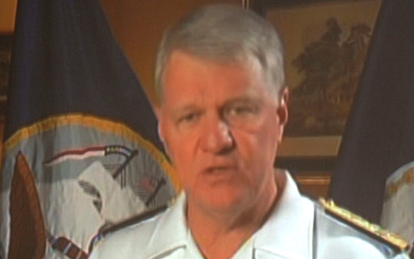 The image shows a projected screen or large display featuring a military officer in a white Navy uniform speaking or giving a briefing. The officer appears to be gesturing with both hands, suggesting an active presentation or address. In the foreground, silhouetted heads of audience members are visible, indicating this is being viewed in a darkened room.