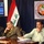 Three men are seated at a conference table in what appears to be an official press conference or meeting for Iraq's Ministry of State Tourism and Antique Affairs. Iraqi flags are displayed prominently in the background alongside the ministry's official seal. A presentation screen displaying Arabic text and contact information is visible to the left.