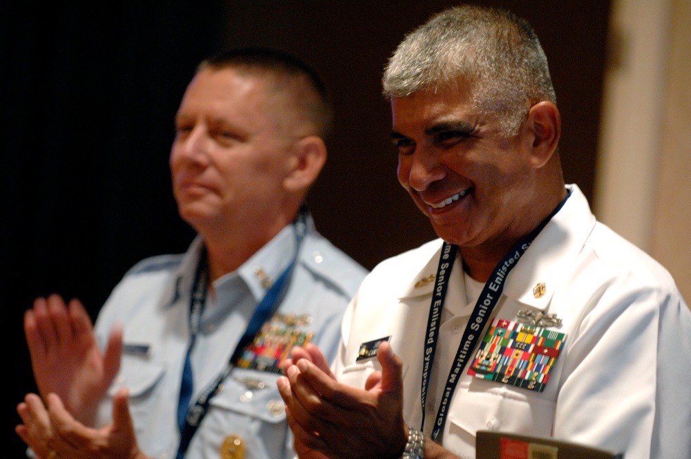 Two military officers in uniform are applauding at what appears to be a formal event or ceremony. The officer in the foreground wears a white naval uniform with extensive medal ribbons and a lanyard reading 'Global Maritime Senior Enlisted.' Both men appear to be in good spirits, with the foreground officer smiling broadly.