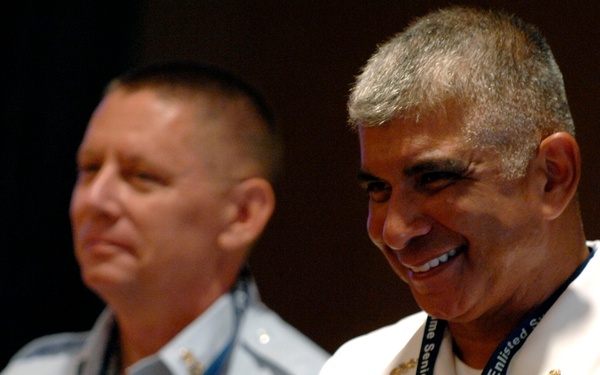 Two military officers in uniform are applauding at what appears to be a formal event or ceremony. The officer in the foreground wears a white naval uniform with extensive medal ribbons and a lanyard reading 'Global Maritime Senior Enlisted.' Both men appear to be in good spirits, with the foreground officer smiling broadly.