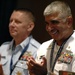 Two military officers in uniform are applauding at what appears to be a formal event or ceremony. The officer in the foreground wears a white naval uniform with extensive medal ribbons and a lanyard reading 'Global Maritime Senior Enlisted.' Both men appear to be in good spirits, with the foreground officer smiling broadly.