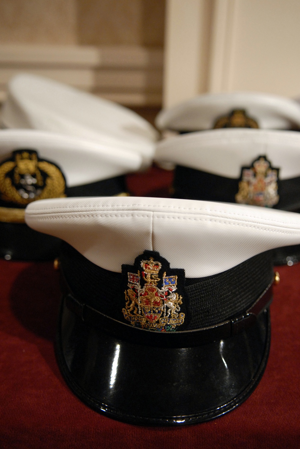 The image shows multiple white-topped naval officer caps with black bands and glossy black peaks arranged on a red surface. Each cap features an embroidered badge depicting what appears to be the British Royal Coat of Arms with a crown, lion, and unicorn motif in gold, red, and blue. The foreground cap is in sharp focus while the background caps are progressively blurred.