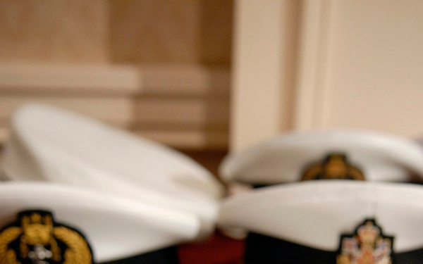 The image shows multiple white-topped naval officer caps with black bands and glossy black peaks arranged on a red surface. Each cap features an embroidered badge depicting what appears to be the British Royal Coat of Arms with a crown, lion, and unicorn motif in gold, red, and blue. The foreground cap is in sharp focus while the background caps are progressively blurred.