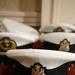 The image shows multiple white-topped naval officer caps with black bands and glossy black peaks arranged on a red surface. Each cap features an embroidered badge depicting what appears to be the British Royal Coat of Arms with a crown, lion, and unicorn motif in gold, red, and blue. The foreground cap is in sharp focus while the background caps are progressively blurred.
