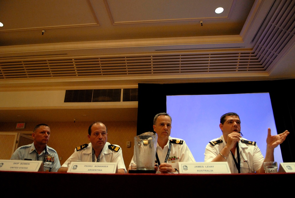 Four military officers from different nations are seated at a panel discussion table in a conference room. Name placards identify them as Skip Bowen (United States), Pedro Bonansea (Argentina), and James Levay (Australia), with a fourth officer partially visible on the far right. The officer on the far right appears to be actively speaking and gesturing with his hand.