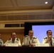 Four military officers from different nations are seated at a panel discussion table in a conference room. Name placards identify them as Skip Bowen (United States), Pedro Bonansea (Argentina), and James Levay (Australia), with a fourth officer partially visible on the far right. The officer on the far right appears to be actively speaking and gesturing with his hand.