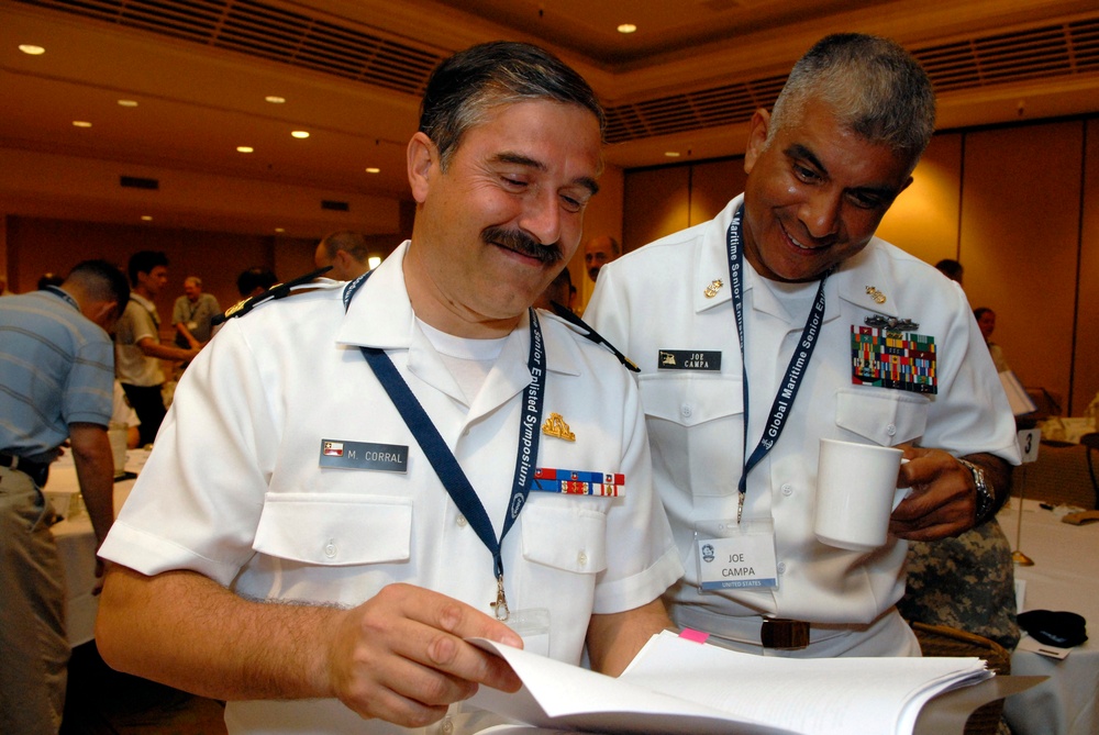 Two military personnel in white dress uniforms are sharing a lighthearted moment while reviewing documents at what appears to be the Global Maritime Senior Enlisted Symposium. The individual on the left is identified by his name tag as M. Corral, and the individual on the right is identified as Joe Campa, United States. Both men are smiling and appear engaged in reviewing printed materials together.