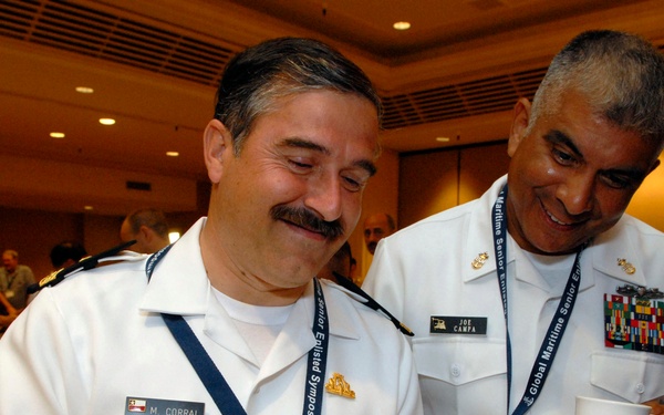 Two military personnel in white dress uniforms are sharing a lighthearted moment while reviewing documents at what appears to be the Global Maritime Senior Enlisted Symposium. The individual on the left is identified by his name tag as M. Corral, and the individual on the right is identified as Joe Campa, United States. Both men are smiling and appear engaged in reviewing printed materials together.