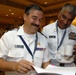 Two military personnel in white dress uniforms are sharing a lighthearted moment while reviewing documents at what appears to be the Global Maritime Senior Enlisted Symposium. The individual on the left is identified by his name tag as M. Corral, and the individual on the right is identified as Joe Campa, United States. Both men are smiling and appear engaged in reviewing printed materials together.