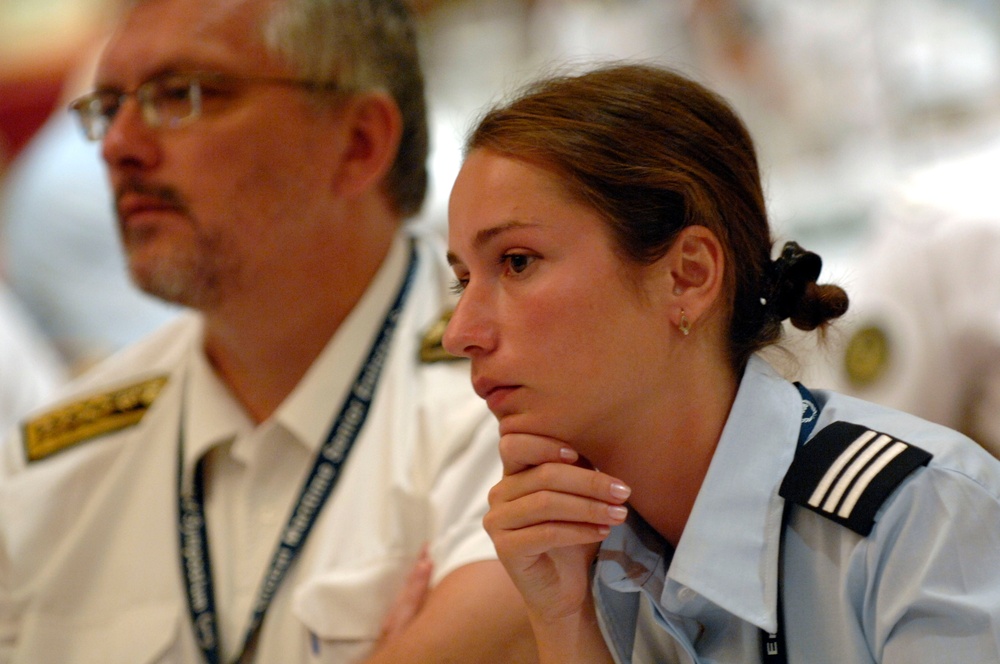Two individuals in uniform are visible, appearing to be attending a formal meeting, briefing, or presentation. The woman in the foreground wears a light blue uniform with black epaulettes bearing three stripes, suggesting a mid-level rank. The man in the background wears a white uniform with gold insignia and a lanyard with an ID badge.