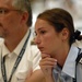 Two individuals in uniform are visible, appearing to be attending a formal meeting, briefing, or presentation. The woman in the foreground wears a light blue uniform with black epaulettes bearing three stripes, suggesting a mid-level rank. The man in the background wears a white uniform with gold insignia and a lanyard with an ID badge.