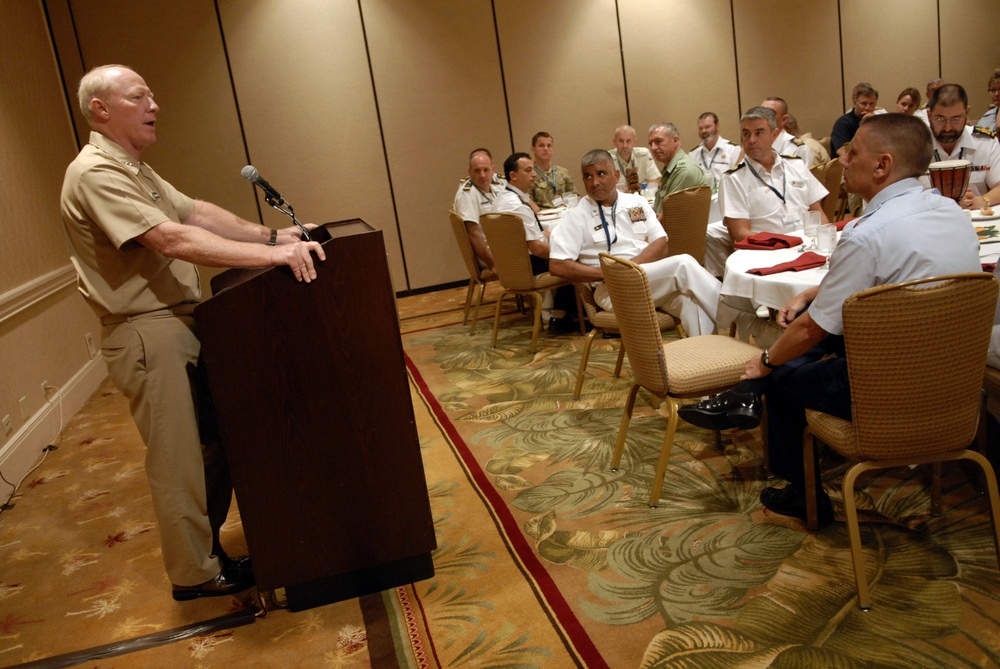 A senior military officer in tan/khaki uniform is delivering a speech at a podium equipped with a microphone to an audience of multinational military personnel seated at round tables in a hotel ballroom or conference room. The audience includes officers in various uniforms including white naval dress uniforms, tan uniforms, and light blue uniforms, suggesting a multinational military conference or symposium. The setting appears to be a formal military dining or briefing event.