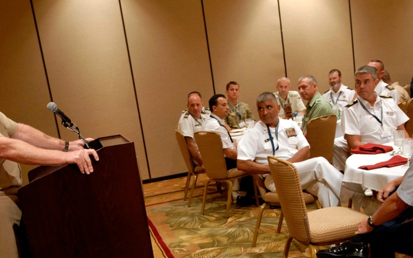 A senior military officer in tan/khaki uniform is delivering a speech at a podium equipped with a microphone to an audience of multinational military personnel seated at round tables in a hotel ballroom or conference room. The audience includes officers in various uniforms including white naval dress uniforms, tan uniforms, and light blue uniforms, suggesting a multinational military conference or symposium. The setting appears to be a formal military dining or briefing event.