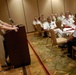 A senior military officer in tan/khaki uniform is delivering a speech at a podium equipped with a microphone to an audience of multinational military personnel seated at round tables in a hotel ballroom or conference room. The audience includes officers in various uniforms including white naval dress uniforms, tan uniforms, and light blue uniforms, suggesting a multinational military conference or symposium. The setting appears to be a formal military dining or briefing event.