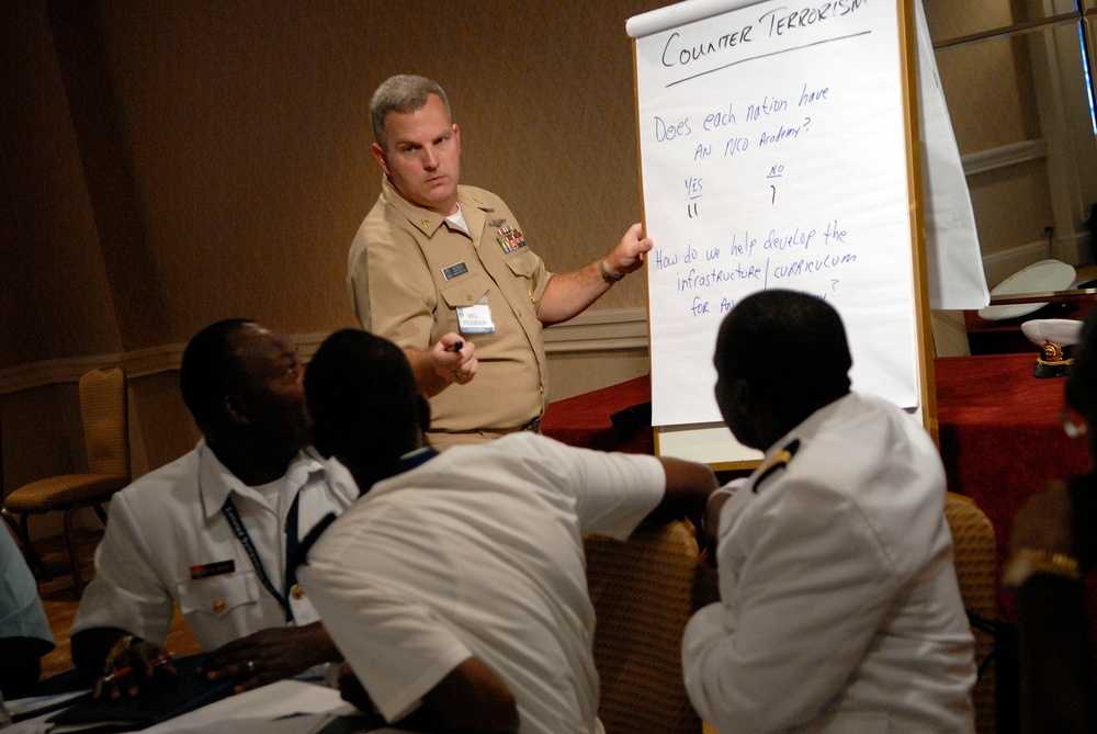A military instructor in U.S. Navy/Marine Corps tan service uniform is conducting a counter-terrorism training session, pointing to a flip chart. The audience consists of several individuals in white naval uniforms, appearing to be international military personnel. The session appears to be a collaborative workshop discussing NCO Academy infrastructure and curriculum development.