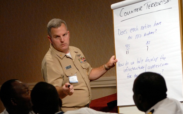 A military instructor in U.S. Navy/Marine Corps tan service uniform is conducting a counter-terrorism training session, pointing to a flip chart. The audience consists of several individuals in white naval uniforms, appearing to be international military personnel. The session appears to be a collaborative workshop discussing NCO Academy infrastructure and curriculum development.