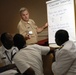 A military instructor in U.S. Navy/Marine Corps tan service uniform is conducting a counter-terrorism training session, pointing to a flip chart. The audience consists of several individuals in white naval uniforms, appearing to be international military personnel. The session appears to be a collaborative workshop discussing NCO Academy infrastructure and curriculum development.