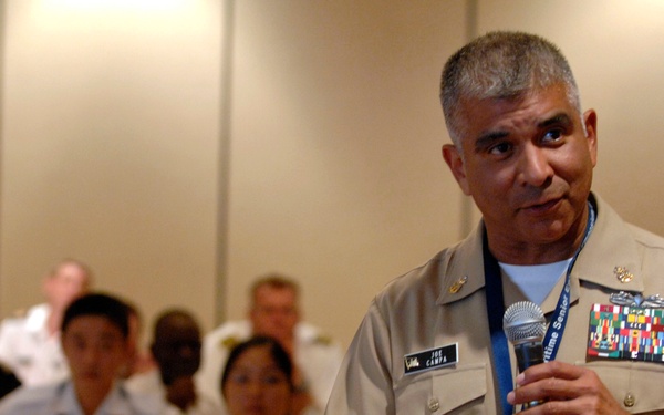 A U.S. Navy officer identified by his name tag as 'Joe Campa' is speaking into a microphone at what appears to be a military conference or seminar. He is wearing a tan/khaki Navy uniform adorned with multiple service ribbons and medals. An audience of military personnel and civilians is seated in the background.