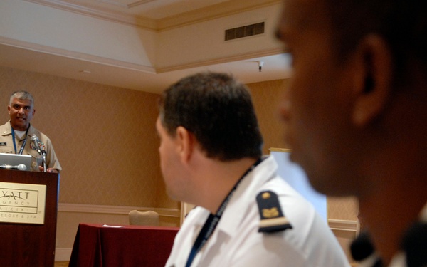 A military or naval officer is presenting at a podium bearing the Hyatt Regency Maui Resort &amp; Spa logo, with a laptop open in front of him. Two uniformed attendees wearing white naval-style uniforms with rank insignia and lanyards are seated in the foreground listening to the presentation. The setting appears to be a formal military or defense conference held in a hotel ballroom.