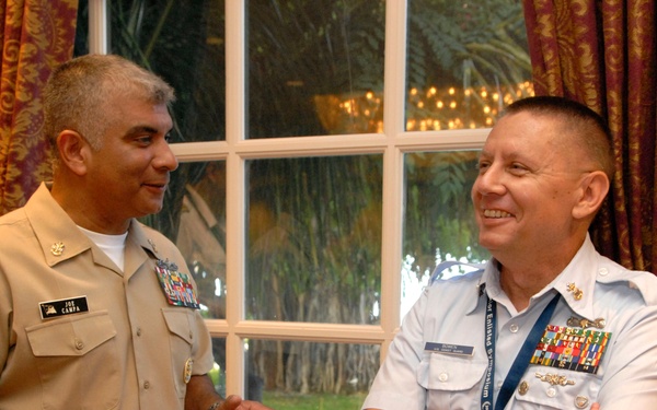 Two military officers in uniform are engaged in a friendly conversation near a window in what appears to be a formal indoor setting. The officer on the left wears a U.S. Navy tan/khaki uniform with a name tag reading 'CAMPA', while the officer on the right wears a light blue uniform consistent with U.S. Air Force dress, with arms crossed and smiling. Both display multiple service ribbons and insignia indicating senior rank.