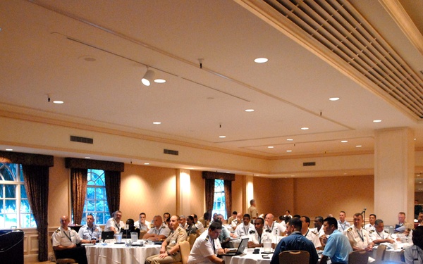 A formal conference or briefing is taking place in a hotel ballroom, with a speaker at a podium addressing a large audience of approximately 40-50 attendees. The majority of attendees appear to be wearing military uniforms of various services and nationalities, suggesting a multinational military conference. A projector is active on the right side of the room, indicating a presentation is in progress.