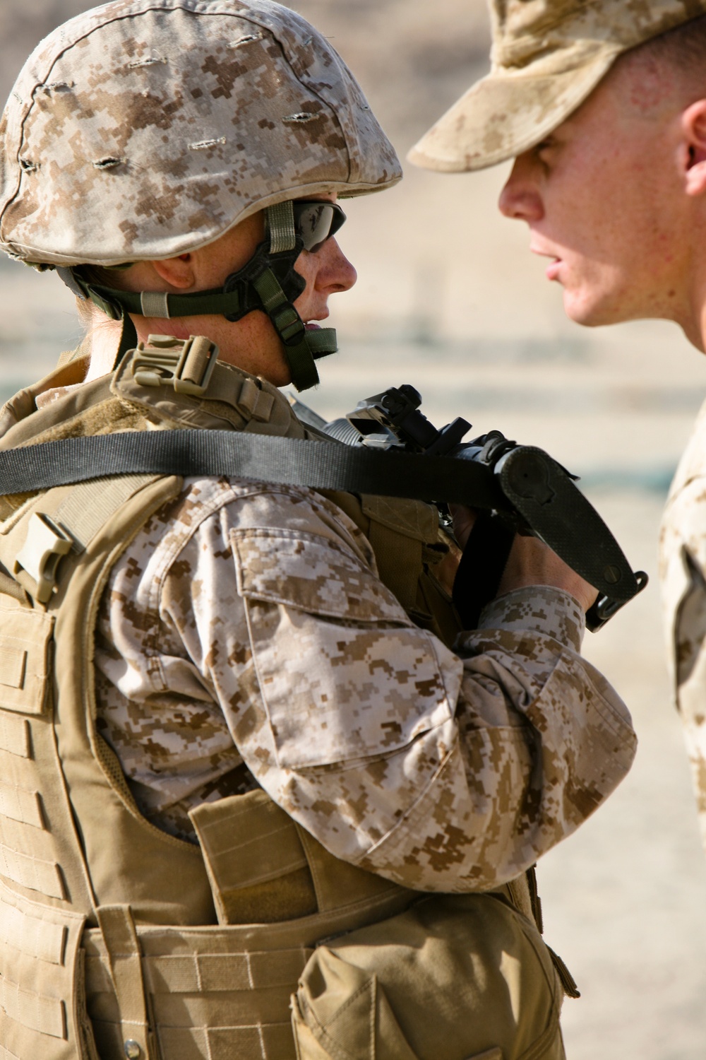 Two military personnel are shown in close proximity, appearing to be in conversation or receiving instruction. Both are wearing MARPAT (Marine Pattern) desert camouflage uniforms, consistent with U.S. Marine Corps equipment. The setting appears to be an arid, outdoor environment.