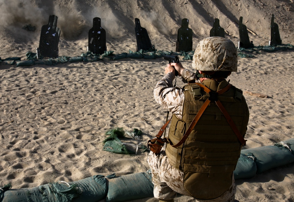 A military service member in desert camouflage (MARPAT pattern) is kneeling on sandbags and firing a pistol at a row of silhouette targets on a sandy shooting range. The shooter is wearing full tactical gear including a plate carrier vest and helmet. Visible dust and debris around the leftmost targets indicate recent impacts from rounds.