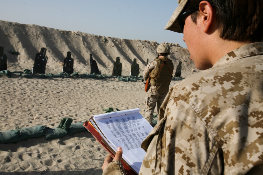 Two military personnel are at an outdoor shooting range in a desert environment. The foreground soldier in MARPAT desert camouflage is reviewing a document or manual, while a second soldier stands downrange near a row of silhouette targets backed by a sand berm. The setting appears to be a training exercise or qualification event.