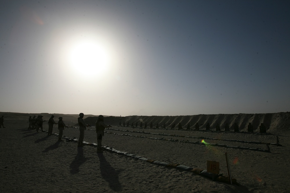 A desert firing range scene photographed into the sun, creating strong silhouette effects. Multiple personnel are positioned along what appears to be a military shooting range with earthen berms serving as target backstops. Items are laid out in rows on the ground in the foreground, consistent with weapons or equipment.