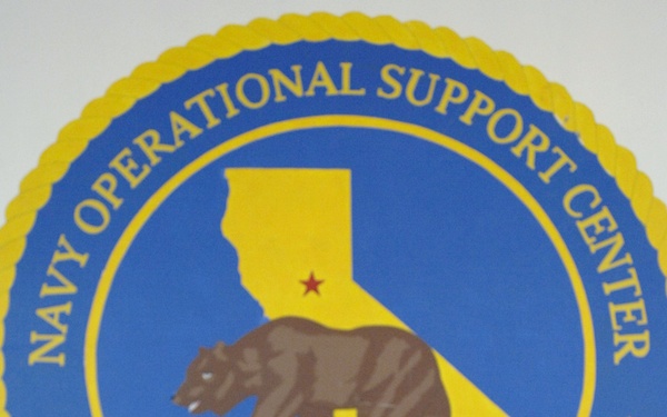 A U.S. Navy officer in tan service uniform is speaking at a microphone while holding a white cup, standing in front of a large Navy Operational Support Center Sacramento seal. The seal features the California state outline with a grizzly bear, consistent with the California state flag imagery. A wooden podium is visible to the left of the speaker.