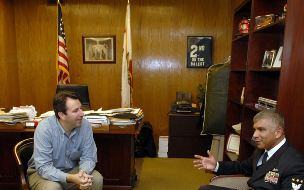 Two men are engaged in conversation in a wood-paneled office setting. One man is in civilian attire seated near a desk, while the other is wearing a dark U.S. Navy senior enlisted dress uniform with gold chevrons and service ribbons. The setting appears to be an official government or congressional office.