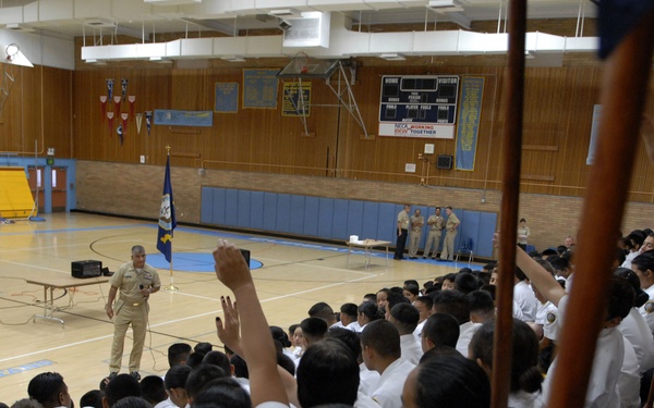 A military or naval officer addresses a large group of cadets or students in uniform inside a gymnasium. The audience, dressed in white uniforms, has several members raising their hands in response to the speaker. The setting appears to be a school or training facility gymnasium with scoreboards, pennants, and flags visible.