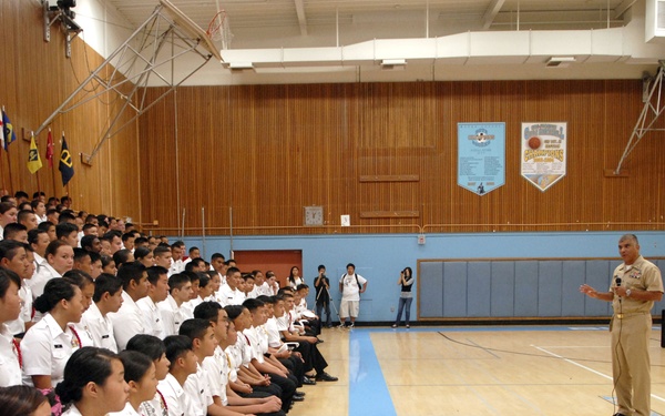 A military or JROTC assembly is taking place in a school gymnasium, where a large group of uniformed cadets are seated in rows listening to a speaker. The speaker, dressed in tan/khaki military uniform, stands at the front holding a microphone addressing the assembled cadets. Several flags are displayed at the back of the gymnasium, and a scoreboard is visible on the right wall.