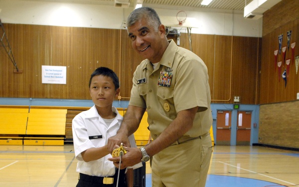 A U.S. Navy officer in tan service uniform and a young student in white uniform are cutting a birthday cake together using a ceremonial sword in what appears to be a school gymnasium. Two large sheet cakes are visible on the table, both decorated with 'Happy Birthday Navy' inscriptions. The setting suggests a Navy birthday celebration event held at a school.
