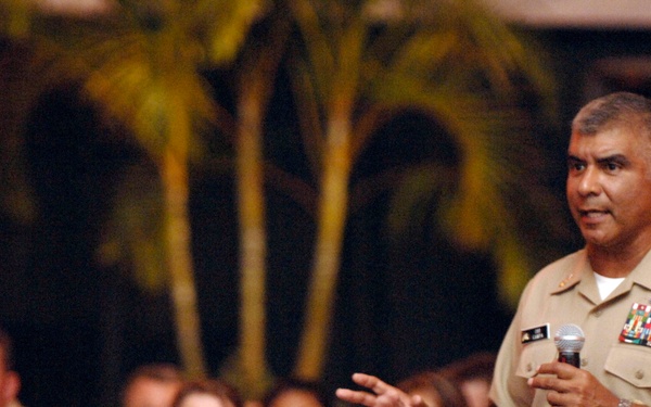 A senior U.S. Navy officer in khaki service uniform is addressing an audience of fellow sailors while holding a microphone. The speaker appears to be delivering a speech or briefing at what looks like an outdoor or semi-outdoor military event. An attentive audience of uniformed Navy personnel is seated in the background.