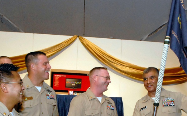 A group of U.S. Navy personnel in tan/khaki service uniforms are gathered at what appears to be an indoor military ceremony or event. One individual on the right is holding a blue military guidon/flag aloft while others laugh and smile. The setting includes decorative gold drapery and tropical plants, suggesting a formal but celebratory occasion.