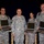 Four U.S. Army soldiers in ACU (Army Combat Uniform) digital camouflage are posed together for a formal photo. Three of the four soldiers are holding open laptop computers, suggesting a technology-related award, presentation, or equipment distribution event. The setting appears to be an indoor venue with a dark blue curtain backdrop and wooden railing.
