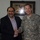 Two men are shaking hands and smiling in what appears to be an indoor meeting setting. One man is wearing a U.S. Army ACU (Army Combat Uniform) with the name tape 'TALLEY' and a single star insignia visible, indicating the rank of Brigadier General. Behind them hangs a framed piece of Arabic calligraphy art on the wall.