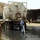 A large white vacuum/suction tanker truck is being operated in a flooded, dark liquid-covered area that appears to be a sewage or wastewater pumping operation. A worker in blue coveralls and a white cap is standing in the liquid while managing a large flexible hose connected to the truck. The scene takes place in what appears to be an industrial or urban area with surrounding buildings and bystanders.