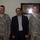 Three men pose for a photo indoors, two wearing U.S. Army ACU (Army Combat Uniform) camouflage uniforms and one civilian in a dark blazer and light shirt. The name tapes visible on the uniforms read 'NEAL' on the left soldier and 'TALLEY' on the right soldier, both bearing 'U.S. ARMY' tape. Behind them hangs a framed piece of Arabic calligraphy art on a light-colored wall.