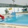 An outdoor swimming pool facility with colorful Arabic murals painted on surrounding white walls. The pool is equipped with a red water slide, a diving board platform, and metal entry ladders. The facility appears newly constructed or recently filled, with a yellow hose still visible on the poolside deck.