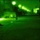 A night vision (green-tinted) image showing an outdoor urban or open area at night. Several human silhouettes are visible standing or moving across what appears to be a open ground or plaza. A row of street lights is visible in the background, illuminating the scene.
