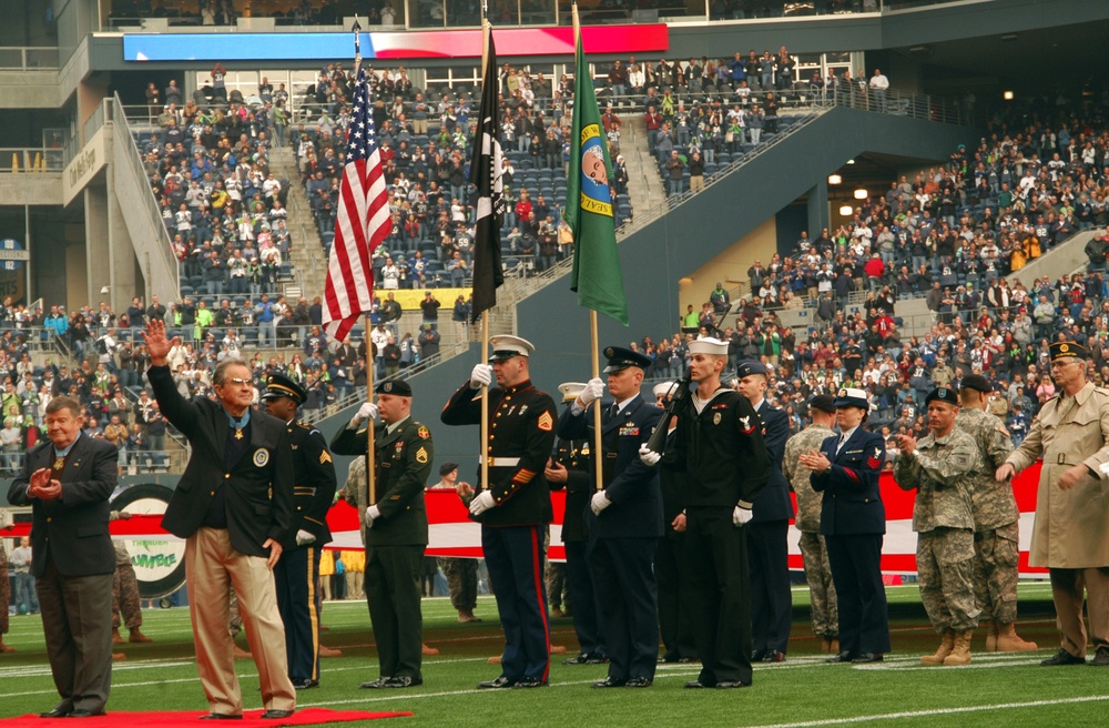 A military color guard ceremony is taking place on the field of a large NFL stadium filled with spectators. Multiple branches of the U.S. military are represented, with service members in dress uniforms holding flags including the American flag, a POW/MIA flag, and what appears to be the Washington State flag. A civilian man wearing what appears to be a Medal of Honor is waving to the crowd.