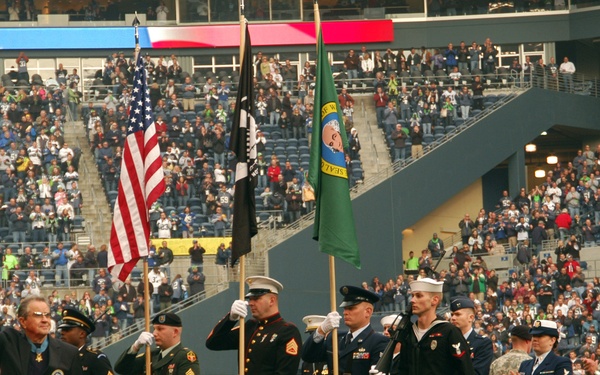 A military color guard ceremony is taking place on the field of a large NFL stadium filled with spectators. Multiple branches of the U.S. military are represented, with service members in dress uniforms holding flags including the American flag, a POW/MIA flag, and what appears to be the Washington State flag. A civilian man wearing what appears to be a Medal of Honor is waving to the crowd.