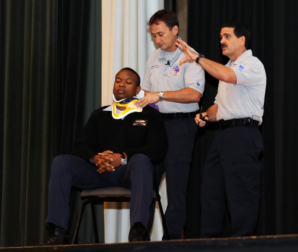Two EMS/paramedic personnel in uniform are demonstrating the application of a cervical collar (neck brace) on a seated young man on what appears to be a stage. This appears to be an educational demonstration or training exercise, likely for a school or public safety awareness event. One paramedic is fitting the collar while the other assists and gestures, possibly explaining the procedure to an audience.