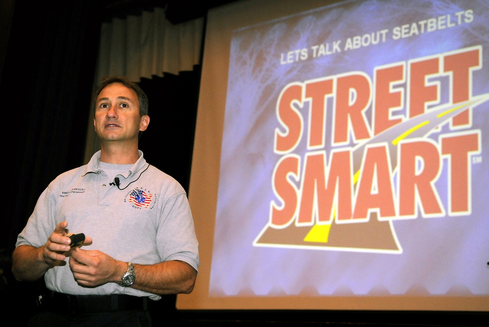 A man is presenting a road safety seminar focused on seatbelt awareness. He stands in front of a projected slide displaying the 'Street Smart' program logo with the heading 'Lets Talk About Seatbelts.' He is wearing a gray polo shirt with an embroidered logo and a lapel microphone, and is holding a presentation clicker.